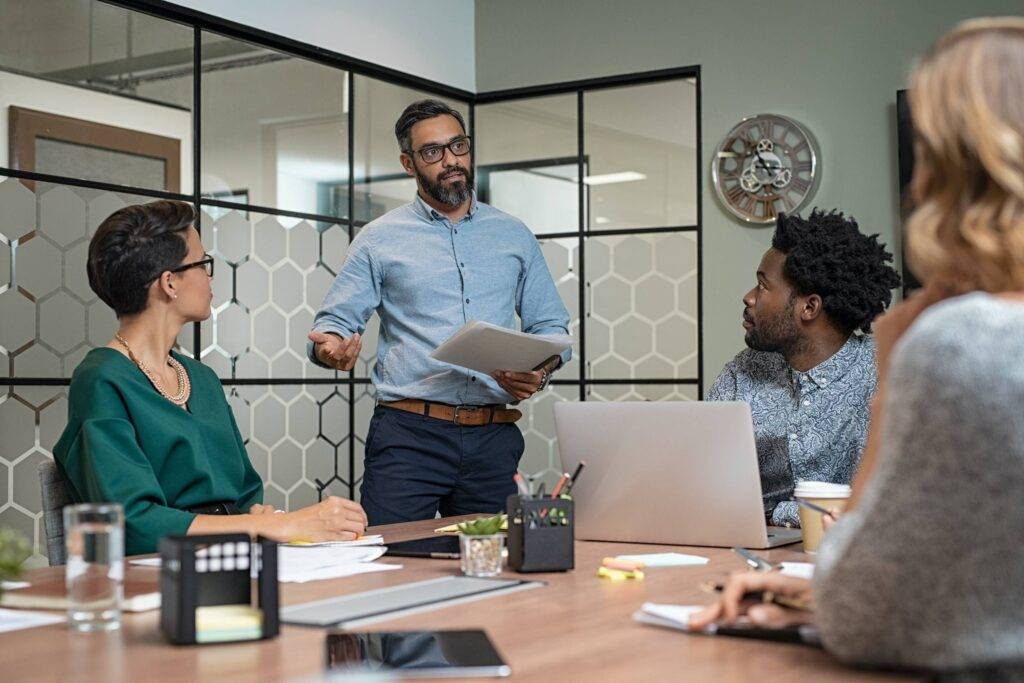 Confident mature businessman giving a presentation to his team in office. Business brief with annual goals with employees and mature boss in meeting room. Multiethnic leader training business people.